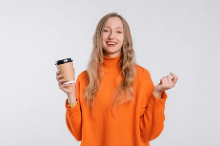 Portrait of a cheerful young woman in an cozy orange sweater holding a paper cup of coffee, standing over neutral backgroundの写真素材