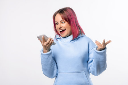 Shocked young woman with pink hair arguing on call, screaming at mobile phone speakerphone, recording voice message with furious face, standing over white backgroundの写真素材