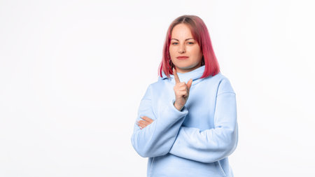 Young woman frowns and shake forefinger, telling no, scolding or telling off someone. Lovely female model with pink hair in a blue hoodie standing over white backgroundの写真素材