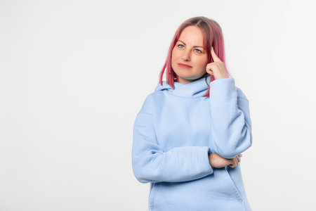 Portrait of young woman with thoughtful face, thinking of something, pondering, has an idea. Lovely female model with pink hair in a blue hoodie standing over white backgroundの写真素材
