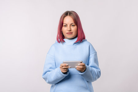 Happy young woman with pink hair playing games on mobile phone, using smartphone, standing in blue hoodie over white backgroundの写真素材