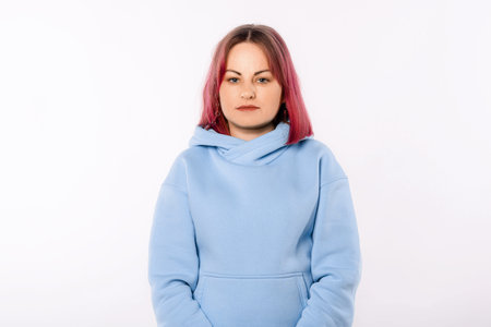 Portrait of young woman with pink hair looking at camera with serious facial expression, wearing a blue hoodie, standing over white backgroundの写真素材