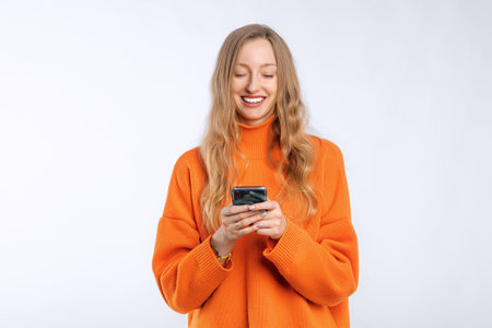 Mobile technology concept. Smiling young woman using his smartphone, holding phone and looking happy, texting in application, standing in an soft orange sweater over white backgroundの写真素材