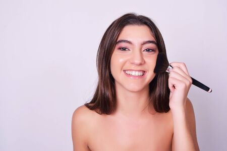 Closeup of young beautiful woman applying foundation powder or blush with makeup brush. Beauty makeup caucasian woman isolated over grey background.の写真素材