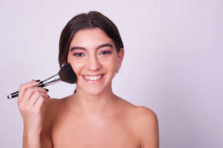Closeup of young beautiful woman applying foundation powder or blush with makeup brush. Beauty makeup caucasian woman isolated over grey background.の写真素材