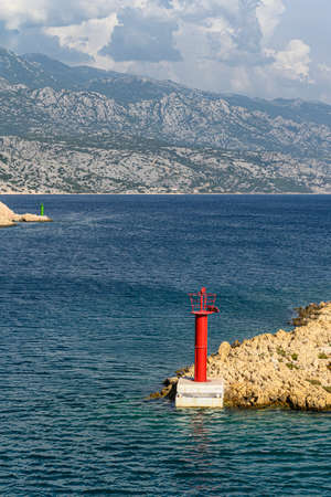 Typical mediterranean red lighthouse on a deserted rocky cape, surrounded by deep blue sea with the storm brewing in the far-away mountains.の写真素材