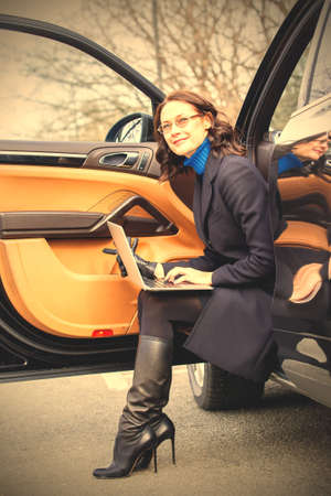 pretty middle-aged woman in a dark coat sitting on the threshold of the car and working on laptop. ...