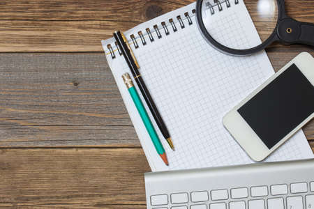 Computer keyboard, notebook, pen, smartphone  and magnifying glass on old wooden table surface....