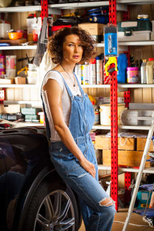 Beautiful curly-haired woman in blue overalls mechanic near a car in the interior of an automobile repair shopの写真素材