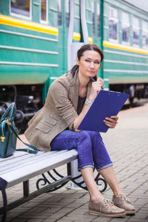 beautiful middle-aged woman sitting on a bench, holding planner and pen in hand and pensive looks away against the background of a passenger train at a railway stationの写真素材