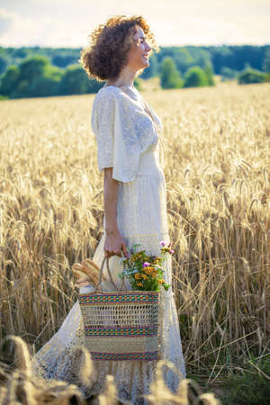 Beautiful smiling Woman Outdoors with wicker bag with natural meal and flowers. Pretty female Model in white dress goes on the summer Field, Sun Light. Glow Sunの写真素材