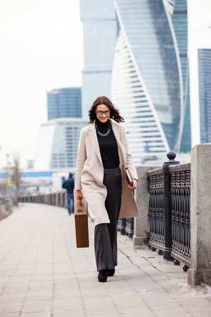 Smiling dark-haired woman in a bright coat is on the waterfront and is in the hands of the case diary and a book. businesswoman goes on the presentation of a new ideasの写真素材