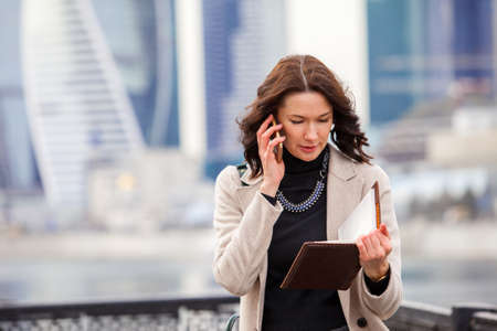 beautiful smiling middle-aged woman using a smartphone with notebook in the business district of a city. businesswoman in a bright coatの写真素材