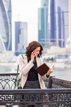 beautiful middle-aged woman in a bright coat with notebook talking on mobile with business partners on a city background. fashion. Business ladyの写真素材