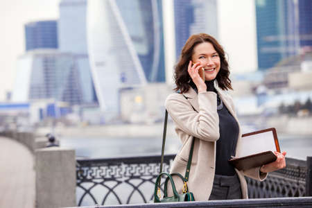 smiling beautiful middle-aged woman using a smartphone in the business district of a city. businesswoman in a bright coatの写真素材