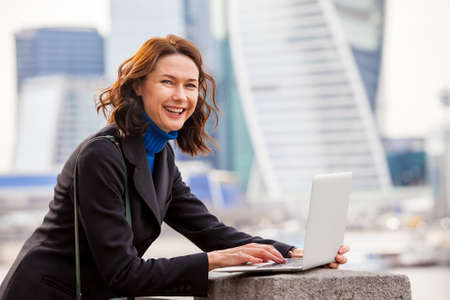 portrait of a beautiful laughing woman with a laptop outdoors with office building behind herの写真素材