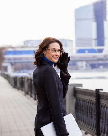friendly brunette with a computer outdoors. woman in a dark coat looked back and smiles. fashion. businessの写真素材
