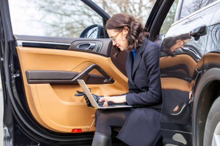 Remote Office Management. Businesswoman in a dark coat sitting on the threshold of the car with the notebook on her lapの写真素材