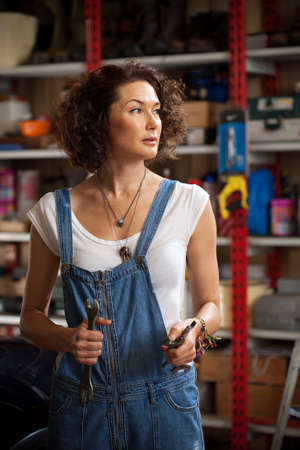 car mechanic woman in blue overalls with a wrench in her hand in the car repair shop. beautiful middle-aged woman with tools to repair the car in the garageの写真素材