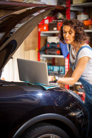 Woman auto mechanic uses a computer to diagnose car engine. Beautiful woman mechanic near a car with an open hood looking at a computer monitorの写真素材