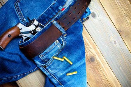 fashion old blue jeans with a leather belt and vintage revolver with cartridges. on a wooden textured backgroundの写真素材