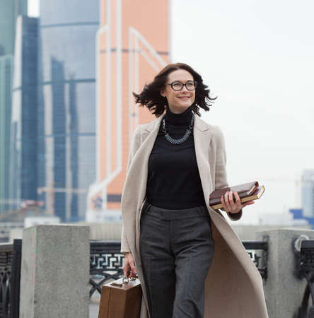 smiling middle aged brunette in a bright coat in the business district of the city. woman with wooden case and books in their hands. fashionの写真素材