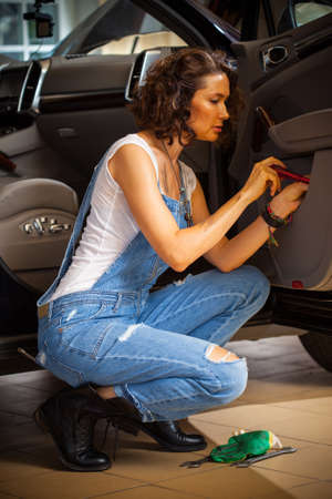 Woman car mechanic in blue overalls adjusts the car door in the workshop. Preparation for autotravelの写真素材