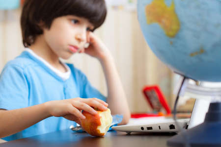 pensive boy sitting at a table with a laptop and eating apple. focus on fruitの写真素材