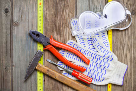 Old hammer, aged pliers, screwdriver, tape measure, gloves and safety glasses on vintage textured boards bench. still life with working toolsの写真素材