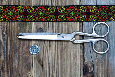 vintage button, emroidered band and a tailor scissors on a textured surface of old boards