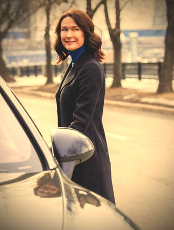 smiling middle aged brunette woman in a dark coat near car outdoors. image filter retro styleの写真素材