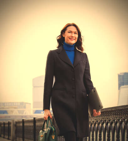 Smiling businesswoman in a dark coat with a laptop and a green hand-bag outdoors.の写真素材