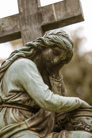 Old gravestone statue of sad thinking woman next to a crossの写真素材