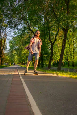 A guy with a beard in a plaid shirt, denim shorts, sunglasses is roller skating at sunset in the city.の写真素材
