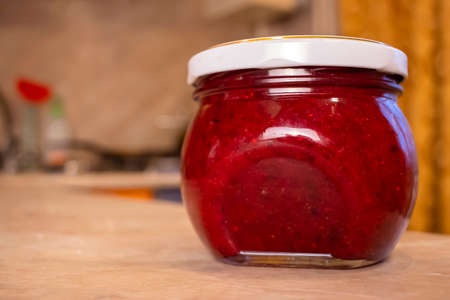 A jar of cranberry jam is on the kitchen table, against the backdrop of an orange curtain. The jar is closed with a white lid with a golden border.の写真素材