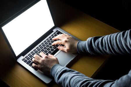 Man working on laptop with white screen glowing in the dark on a wooden desk seen from over the...