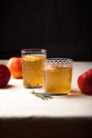 Two vintage glasses with apple cider on black background. Christmas beverages concept. Two red apples and rosemary sprig aside.  Warm backlight. Vertical composition.の写真素材