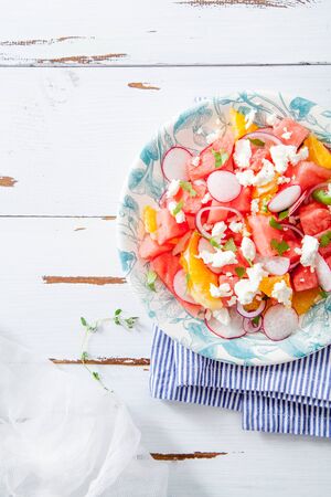 Watermelon salad with orange, radish and red onion on white distressed wooden table. Summer raw organic food concept. Close-up. の写真素材