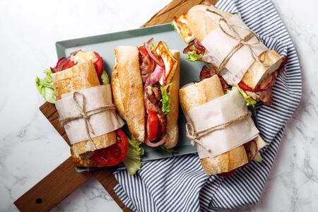 Fresh baguette sandwich bahn-mi styled. Bacon, roasted cheese, tomatoes and lettuce on metallic tray on white marble background.の写真素材