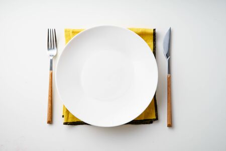 White empty plate with knife and fork and yellow textile napkin on white background
