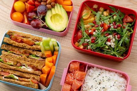 Creative layout with healthy lunch dishes variety in bento boxes on wooden table. Sandwich, slad with grains and pomegranate seeds, salmon with rice and fruit snacks. Daily office or school lunch conceptの写真素材