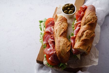 Top view of two fresh baguette sandwiches bahn-mi styled. Ham, sliced cheese, tomatoes and fresh lettuce on dark wooden cutting board on concrete background.の写真素材