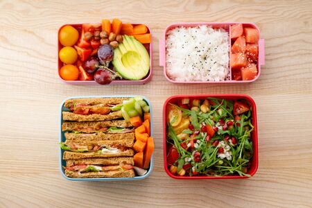 Creative layout with healthy lunch dishes variety in bento boxes on wooden table. Sandwich, slad with grains and pomegranate seeds, salmon with rice and fruit snacks. Office or school lunch conceptの写真素材