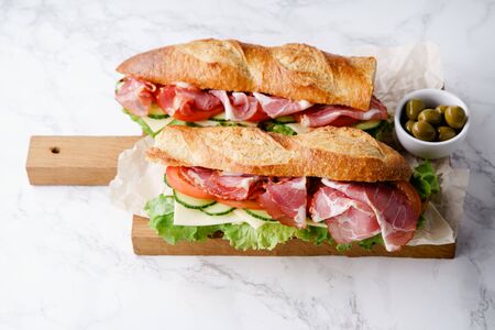 Fresh baguette sandwich bahn-mi styled. Ham, sliced cheese, tomatoes and fresh lettuce on wooden cutting board on white marble background.の写真素材