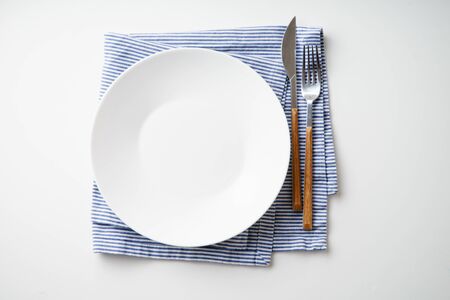 White empty plate with knife and fork on striped blue and white textile napkin on white backgroundの写真素材