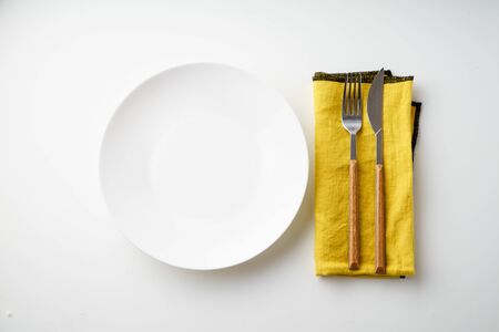 White empty plate with knife and fork and yellow textile napkin on white background