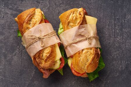 Fresh big baguette sandwich with bacon, chedder cheese, mustard, lettuce and vegetables on dark backgroundの写真素材