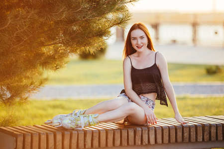 woman in roller skates sitting on a bench on it wearing a blue shirt and light in the form of chert skating dresses prohodile in the city park around the growing firの写真素材