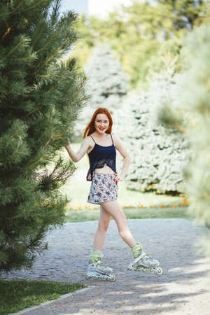 red-haired woman in roller skate in a city park on her T-shirt and bright blue chert in the park grow spruce lined road pavementの写真素材