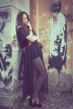 portrait of a beautiful young sad goth girl in a black dress standing in an abandoned old building outdoors at the day timeの写真素材